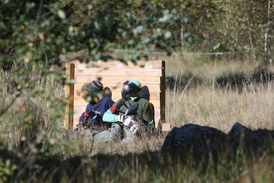 Två personer som spelar paintball sitter bakom ett träplank och tar skydd.